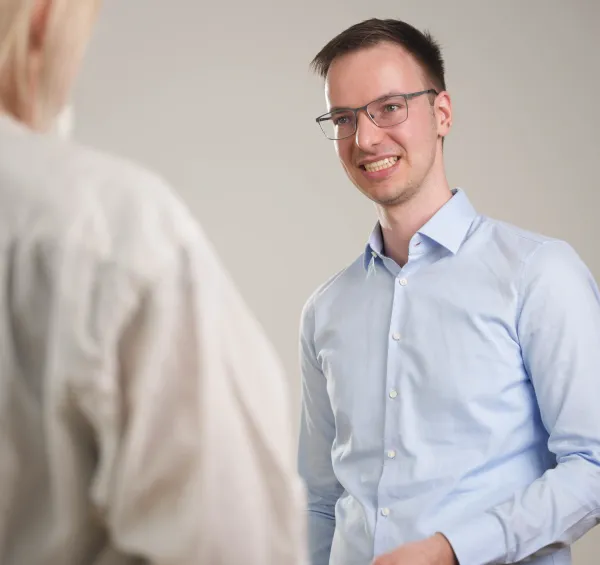 Junger Geschäftsmann in hellblauem Hemd und Brille lächelt während eines Gesprächs mit einer Person im Vordergrund, die teilweise von hinten zu sehen ist. Professionelle Büroatmosphäre vor hellem Hintergrund.
