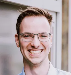 Nahaufnahme eines jungen Mannes mit Brille, der ein strahlendes Lächeln zeigt. Er trägt ein hellblaues Hemd, hat kurzes braunes Haar und wird vor einem hellen, unscharfen Hintergrund fotografiert.