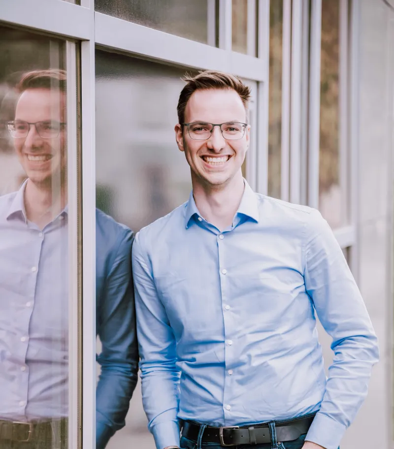 Junger Mann in hellblauem Businesshemd und Brille lehnt an einer Glasfassade, wobei sein Spiegelbild in der Scheibe zu sehen ist. Er trägt einen schwarzen Gürtel und lächelt freundlich in die Kamera. Die Aufnahme hat eine helle, professionelle Atmosphäre.